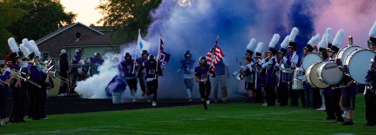 football players running through smoke with band holding instruments